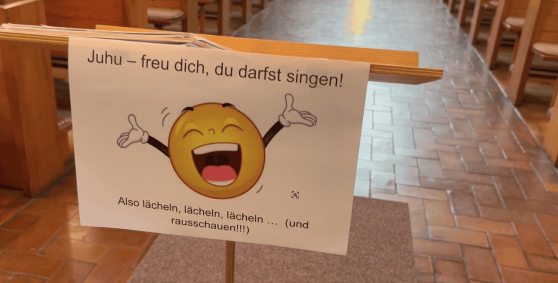 Ein aufgestelltes Schild auf der Kirchenbank mit der Aufschrift „Juhu – freu dich, du darfst singen! Also lächeln, lächeln, lächeln … und rausschauen!“ neben einem lachenden Smiley