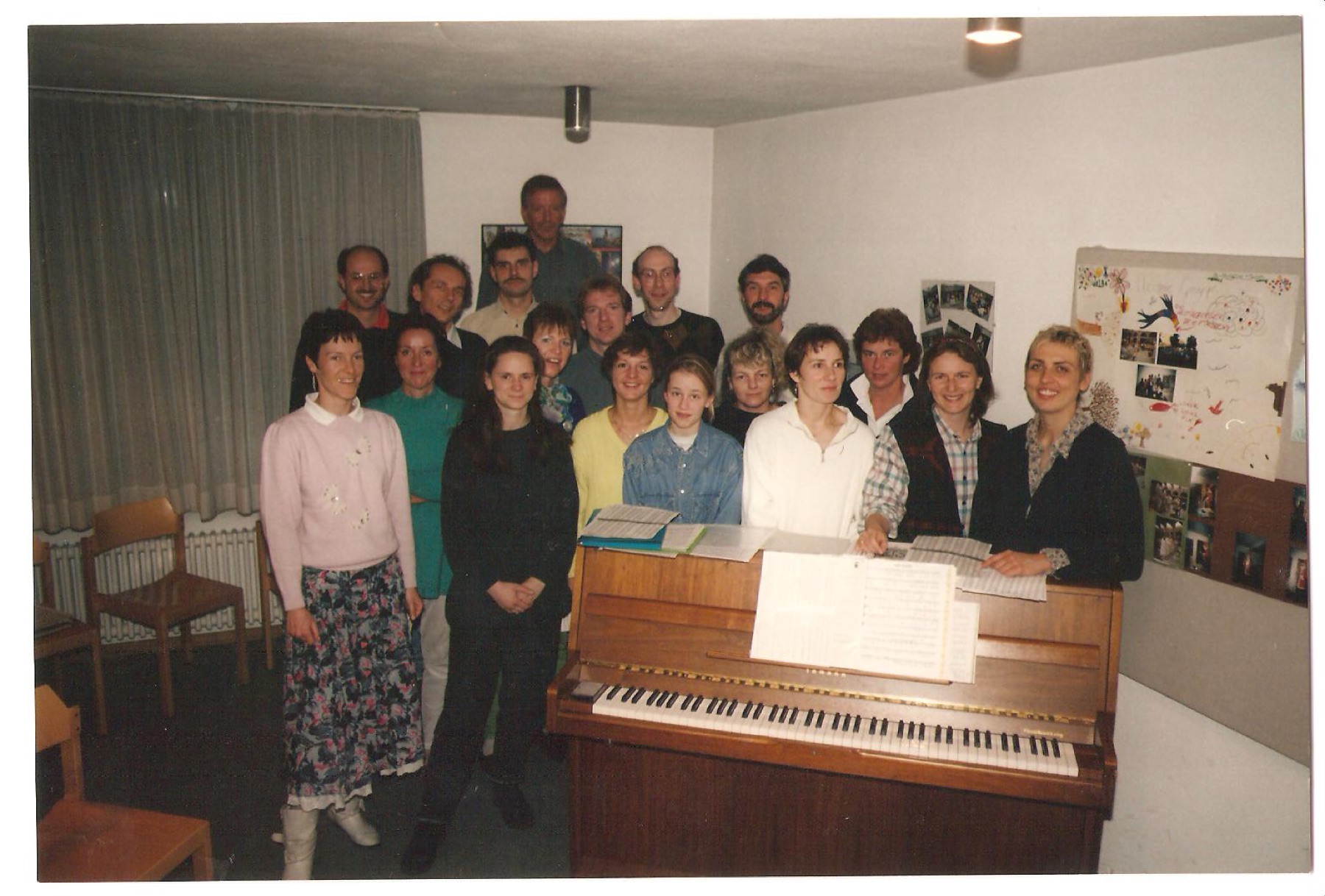 Der Chor im Jahr 1996 im Probenraum von St. Rita: rund zwanzig Sängerinnen und Sänger stehen um ein Klavier mit aufgeschlagenen Noten